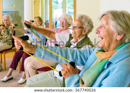 Large group of happy enthusiastic elderly ladies exercising in a gym sitting in chairs doing stretching exercises with rubber bands