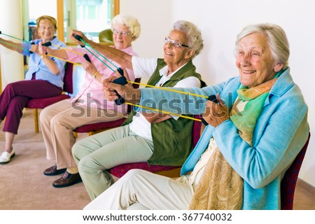 Group of four smiling senior women toning their arms with elastic strengthening bands while seated in fitness class