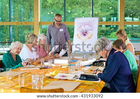 Group of six female senior citizens at long table in painting class with male teacher in glasses standing near front