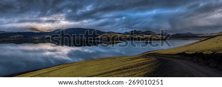 Panorama of overcast sky over mountains reflected in lake at daybreak with a winding road in the foreground, Landmannalaugar, Iceland.