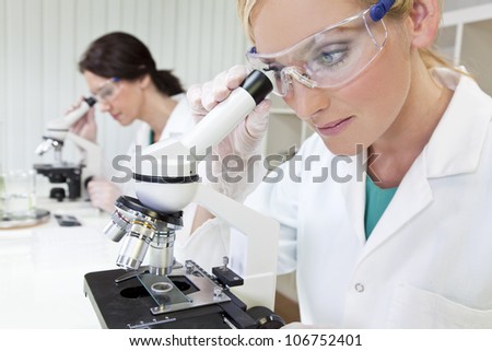 A blond female medical or scientific researcher or doctor using her microscope in a laboratory with her colleague out of focus behind her.