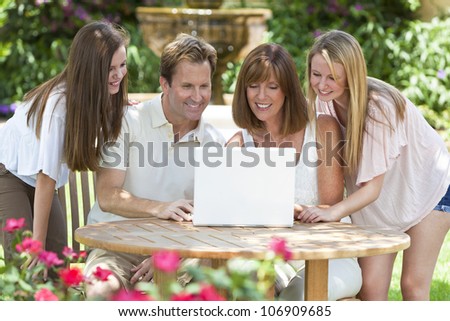 An attractive happy, smiling family of mother, father and two daughters sitting using a laptop computer outside in the garden in warm summer sunshine