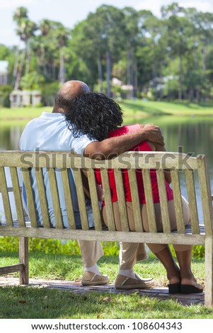 Rear view of a happy romantic senior African American couple sitting on a park bench embracing looking at a lake