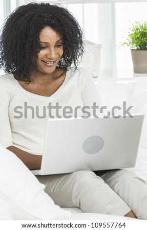 Beautiful smiling African American woman at home sitting on sofa or settee using her laptop computer.