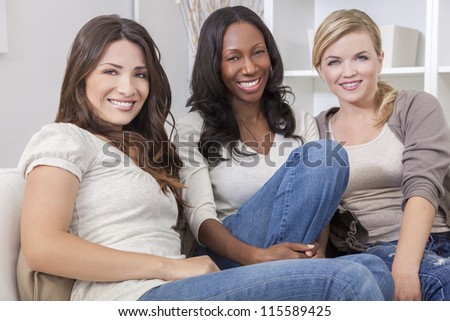 Interracial group of three beautiful young women friends at home sitting together on a sofa smiling and having fun