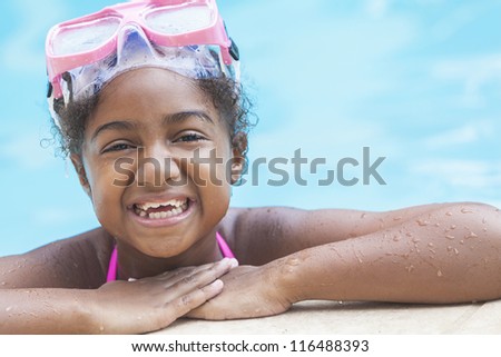 A cute happy young African American girl child relaxing on the side of a swimming pool smiling & wearing pink goggles