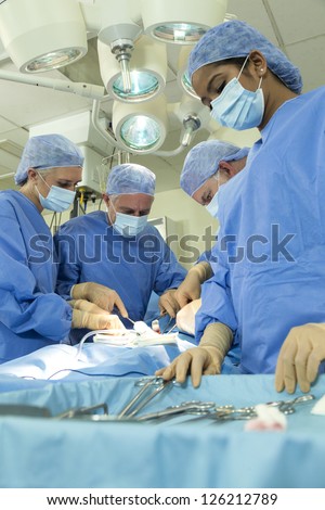 A team of interracial medical doctors male & female surgeons in surgery operating on a patient using different equipment & instruments wearing scrubs and masks