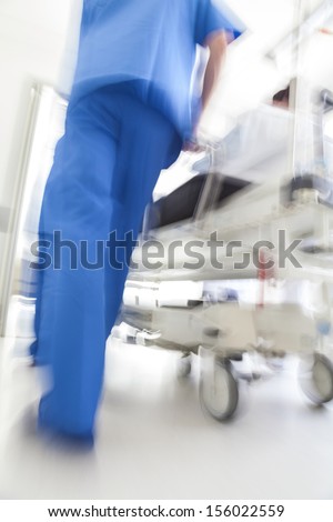 A Motion Blurred Photograph Of A Patient On Stretcher Or Gurney Bed Being Pushed At Speed Through A Hospital Corridor By Doctors &Amp; Nurses To An Emergency Room