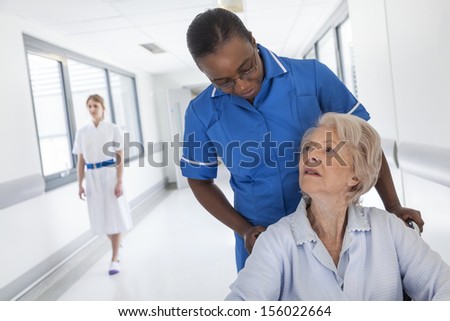 Senior female woman patient in wheelchair being pushed down hospital corridor with African American female nurse