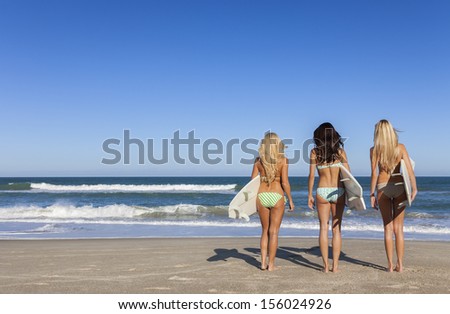 Rear view of three Beautiful young women surfer girls in bikinis with white surfbords at a beach
