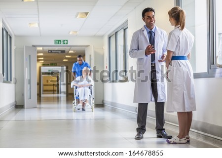 Asian Indian Male Doctor &Amp; Female Nurse In Hospital Corridor With A &Amp; Senior Female Wheelchair Patient And African American Nurse In Background