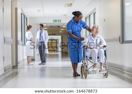 Senior Female Woman Patient In Wheelchair Sitting In Hospital Corridor With African American Female Nurse Doctor And Nurse In Background