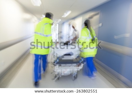 A motion blurred photograph of a patient on stretcher or gurney being pushed at speed through a hospital corridor by doctors & nurses to an accident and emergency room