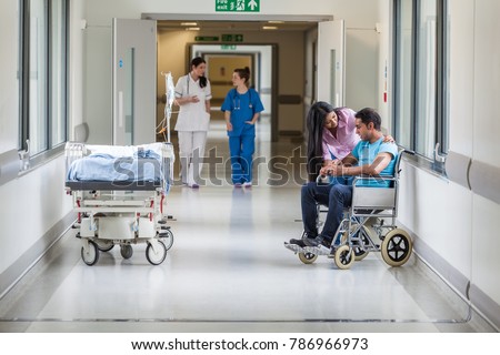 Male Asian Indian wheelchair patient with his wife or girlfriend & nurses in hospital corridor