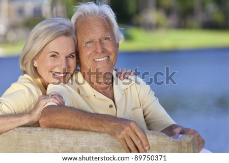 Happy smiling romantic senior man and woman couple sitting on a park bench embracing by a blue lake