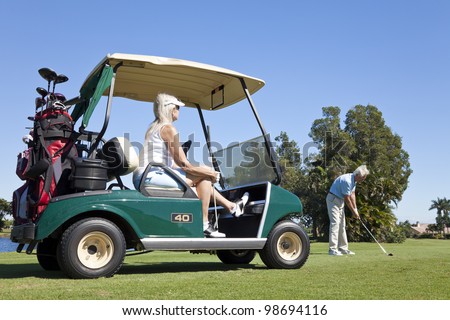 Happy senior man and woman couple together playing golf, the man is hitting a shot the woman is sitting in a golf cart