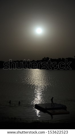 People silhouette swimming and playing in lake water and a floating platform. Full moon in the sky and water trail reflection in the water.