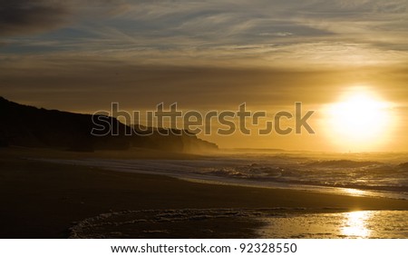 Rock cliffs and beach ocean waves against a full sun sunset. Foz do Arelho, Portugal.