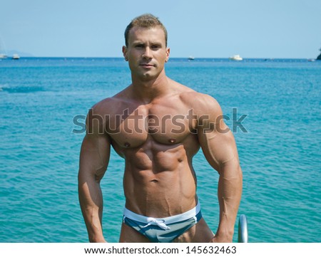Handsome young bodybuilder standing with sea or ocean behind showing muscular torso, pecs, arms and abs