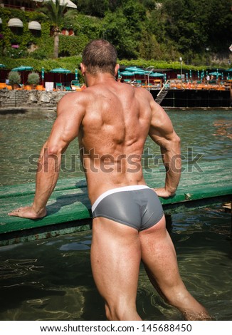 Handsome young bodybuilder\'s back with sea or ocean behind showing muscular legs, buttocks and shoulders