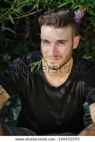 Attractive, smiling young man outdoors with flower between his lips, looking in camera