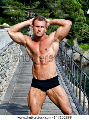 Attractive young muscle man smiling, outdoors, showing muscular body with hands behind his head