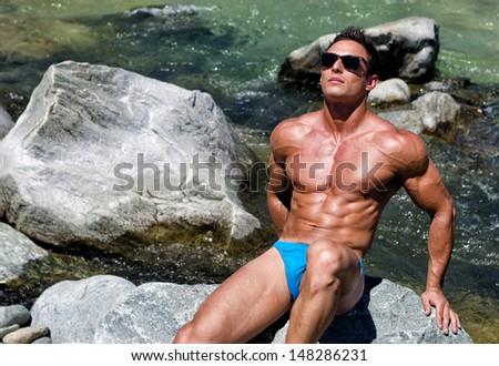 Attractive, tanned young muscle man sitting on stone by river water, in the sun with sunglasses
