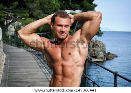 Attractive young muscle man smiling, outdoors, showing muscular body with hands behind his head