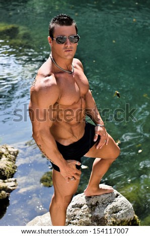 Handsome young muscle man standing in water pond shirtless with sunglasses