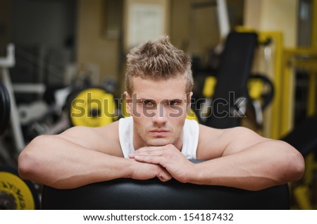 Handsome young man in gym resting on gym equipment, looking in camera