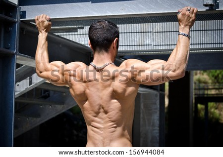 Muscular back of shirtless male bodybuilder hanging from metal structure outdoors