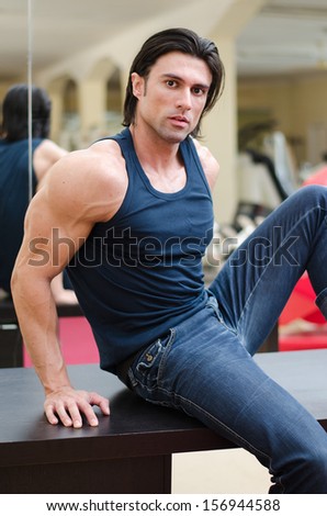 Attractive, muscular man sitting on desk indoors, wearing jeans and tanktop