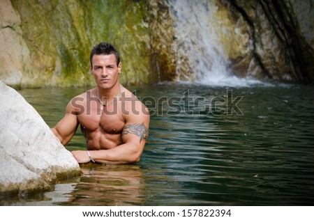 Handsome young muscle man standing in water pond, with waterfall behind