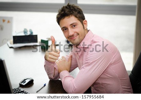 Cheerful office worker at desk doing thumb up sign and smiling. Real workplace