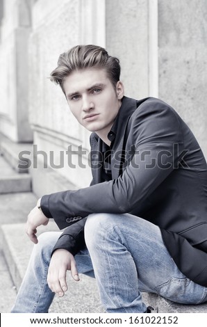 Attractive blond young man in jeans and jacket, sitting outdoors looking in camera