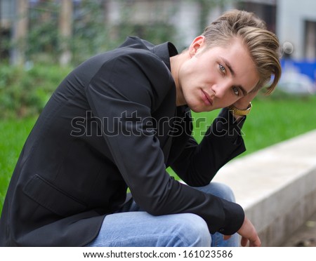 Serious or sad blond young man in jeans and jacket, sitting outdoors looking in camera