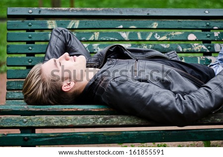 Handsome blond young man lying down on green, wooden park bench, looking up