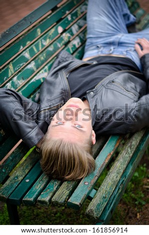 Handsome blond young man lying down on green, wooden park bench, looking at camera