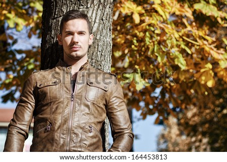 Handsome young man against tree in autumn, wearing leather jacket
