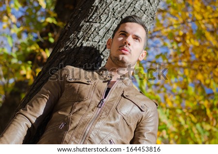 Handsome young man against tree in autumn, wearing leather jacket