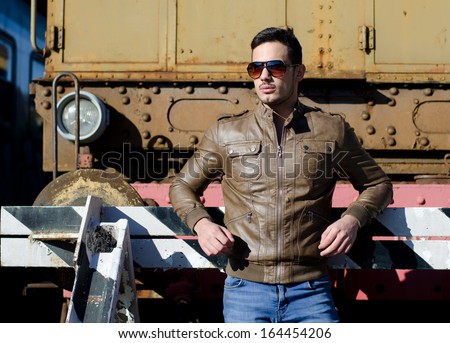 Attractive young man in leather jacket and jeans in front of old train wearing sunglasses