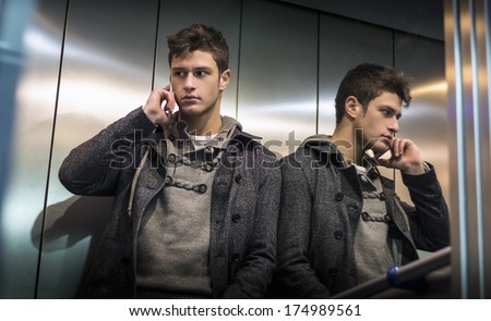 Handsome young man in elevator (lift) using cell phone, his reflection on mirror