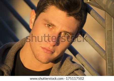 Headshot of handsome young man, lit by sunset light, leaning against handrail