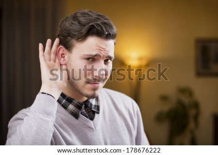 Handsome young man can\'t hear, putting hand around his ear. Indoors shot inside a house