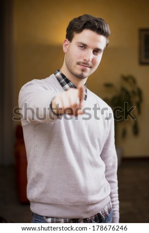 Handsome young man pointing finger at you, smiling. Indoors shot inside a house