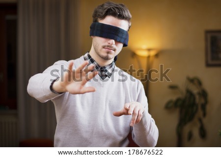Blindfolded young man at home in living room cannot see, trying to find his way with his hands