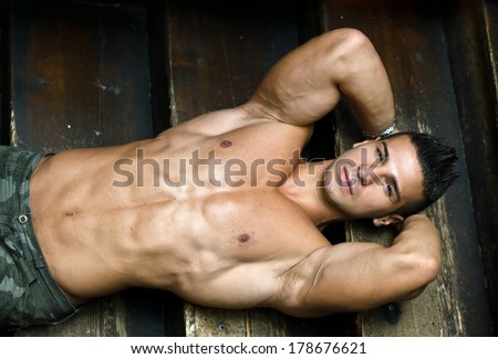 Attractive muscular young man laying on steps of wooden stair, looking at camera, shirtless