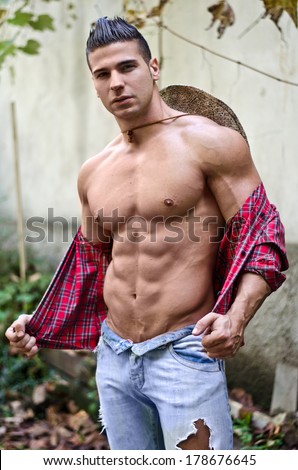 Handsome, muscular young man taking off shirt, wearing jeans and straw hat, outdoors