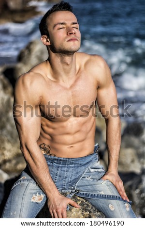 Muscular shirtless young man sunbathing on rock by the sea or ocean, eyes closed
