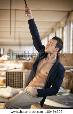 Muscular young man in messy workplace, hanging from tube on ceiling, looking up